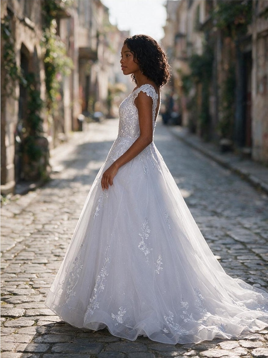 Lacey Bridal Ltd. Seraphina A-line wedding dress with floral lace, cap sleeves, and flowing tulle on a cobblestone street