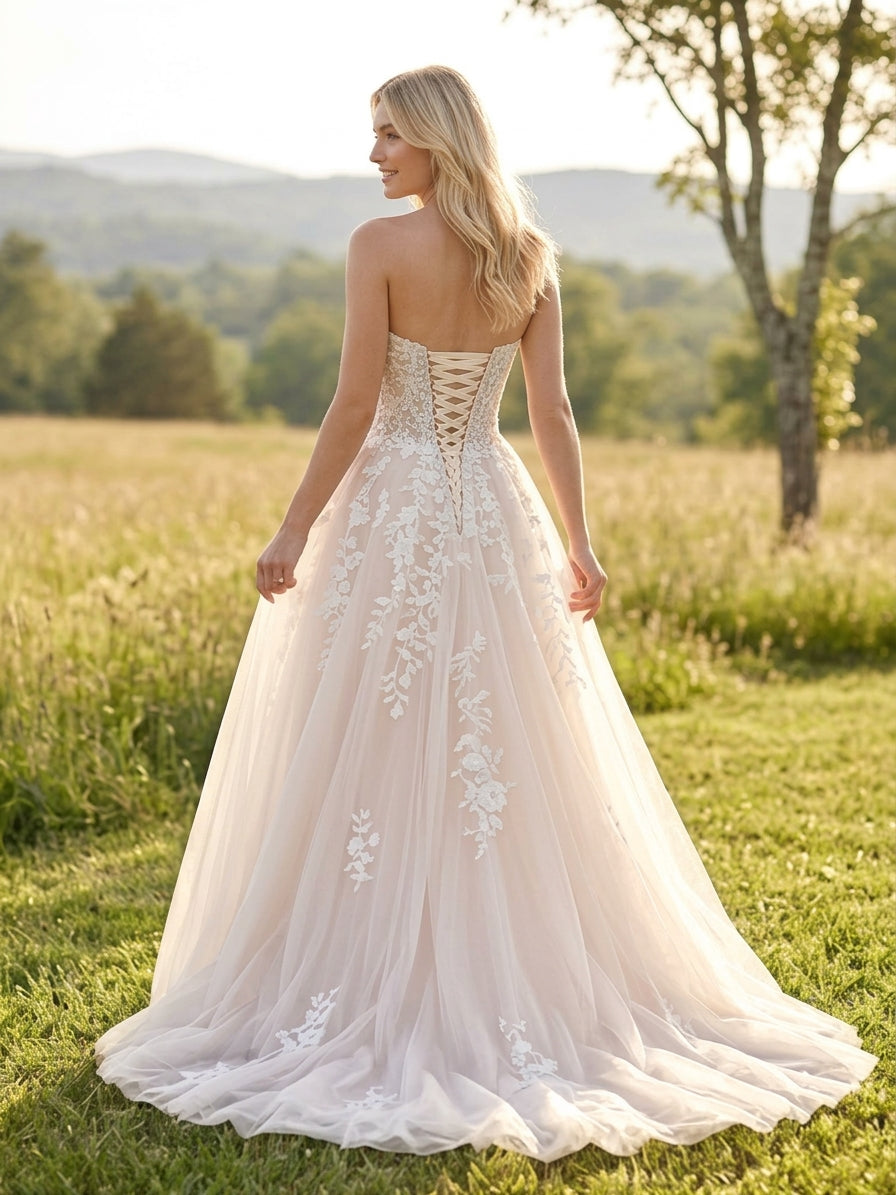 Lacey Bridal Ltd. Sibylla blush A-line wedding gown with floral lace appliqué, corset back, and tulle skirt in a field.